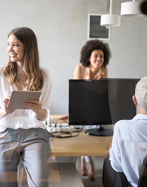 Jeune femme souriante avec tablette dans un bureau moderne, entourée de collègues en pleine collaboration.